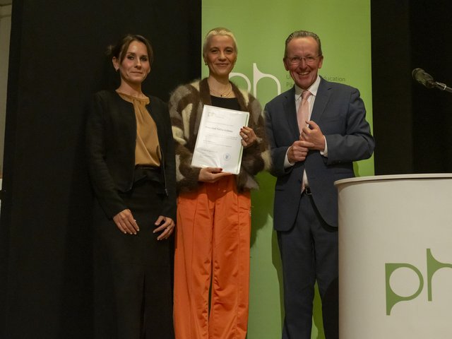 Drei Personen stehen auf einer Bühne vor einem grünen Hintergrund, die Person in der Mitte hält eine Urkunde und lächelt. Rechts daneben steht ein Mann im Anzug neben einem Rednerpult, während die Person links daneben ruhig in die Kamera blickt.