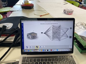 Ein Laptop zeigt ein neuronales Netzwerk Diagramm und einen kleinen Roboter; auf dem Tisch im Hintergrund sind zwei ähnliche Roboter, Papiere und Stifte.