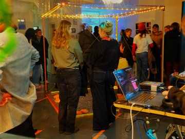 Situation auf der Kunstausstellung mit ein paar Personen, die eine Installation bettrachten, und weiteren im Hintergrund.