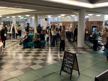 Im Foyer der Stadtbibliothek Karlsruhe treffen TeilnehmerInnen zum Fachtag für Lesepat:innen und Lehrkräfte ein. Es sind Personen im Gespräch zu sehen.