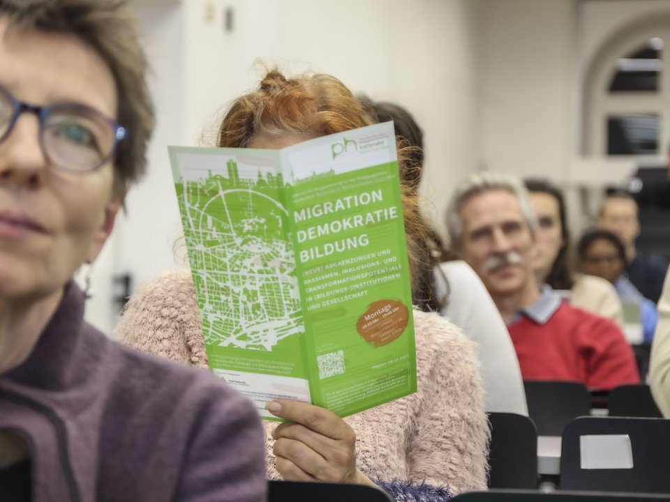 Im Publikum hebt jemand einen Flyer mit dem Titel "Migration Bildung Demokratie" hoch