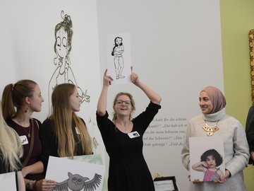 Eine junge Frau hält eine Zeichnung von einer Frau im Comic-Stil in die Höhe und erklärt etwas, drei weitere Frauen hören ihr zu.