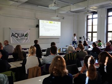 Eine Person hält einen Vortrag. Im Vordergrund sitzen Zuhörende.