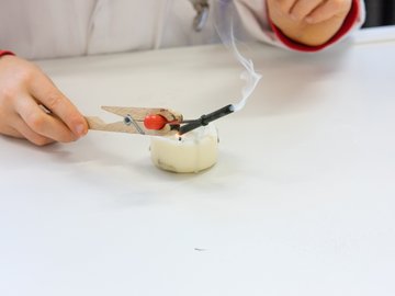 Ein Kind experimentiert mit einer brennenden Kerze und einem Röhrchen, das per Klammer über die Flamme gehalten wird. 