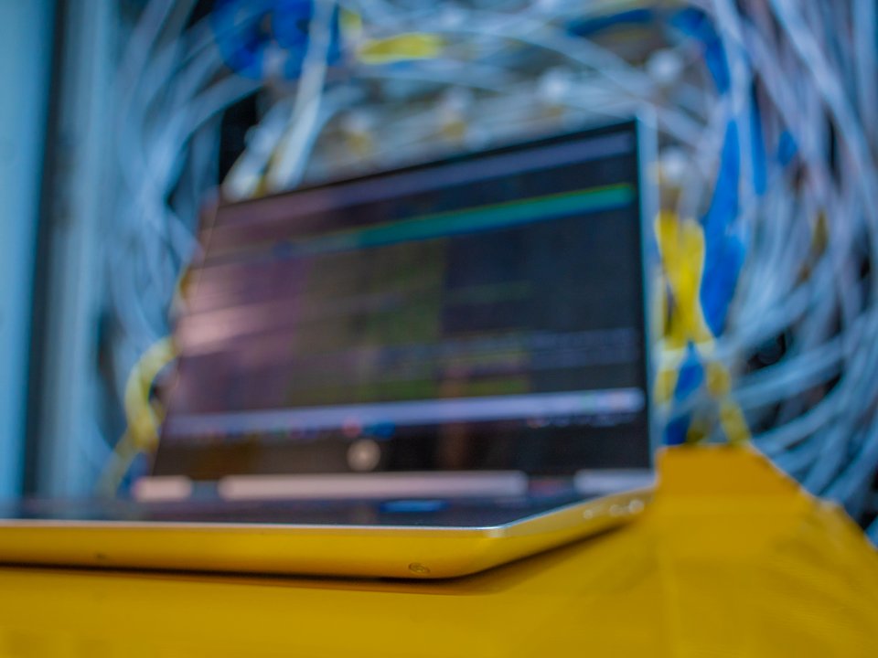 Unscharfes Foto eines Laptops auf einer gelben Oberfläche, im Hintergrund verlaufen zahlreiche Netzwerkkabel in Blau und Weiß, was auf eine Server- oder IT-Umgebung hindeutet.