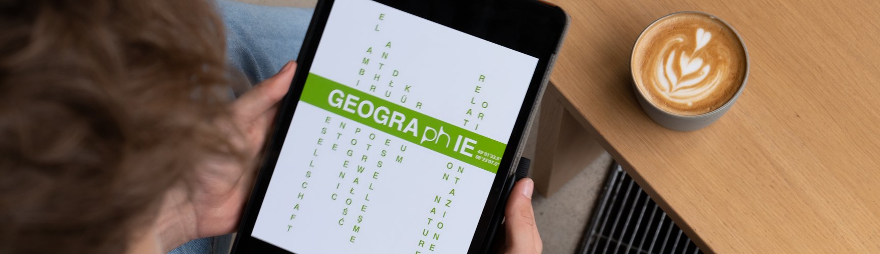 Es wird ein Tablet mit dem Logo des Faches gezeigt. Das Logo zeigt die Bedeutung von Geographie an der Pädagogischen Hochschule Karlsruhe, wie Raum mehrsprachig zwischen Gesellschaft und Umwelt verhandeln.