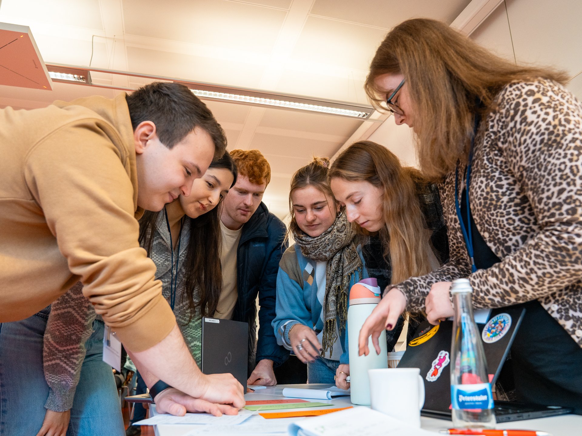 Eine Gruppe Studierender im Austausch um einen Tisch herum stehend.