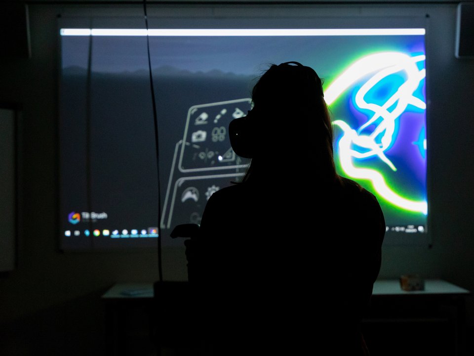 Eine Person mit einer VR Brille steht in einem dunklen Raum und schaut auf einen erleuchteten Monitor