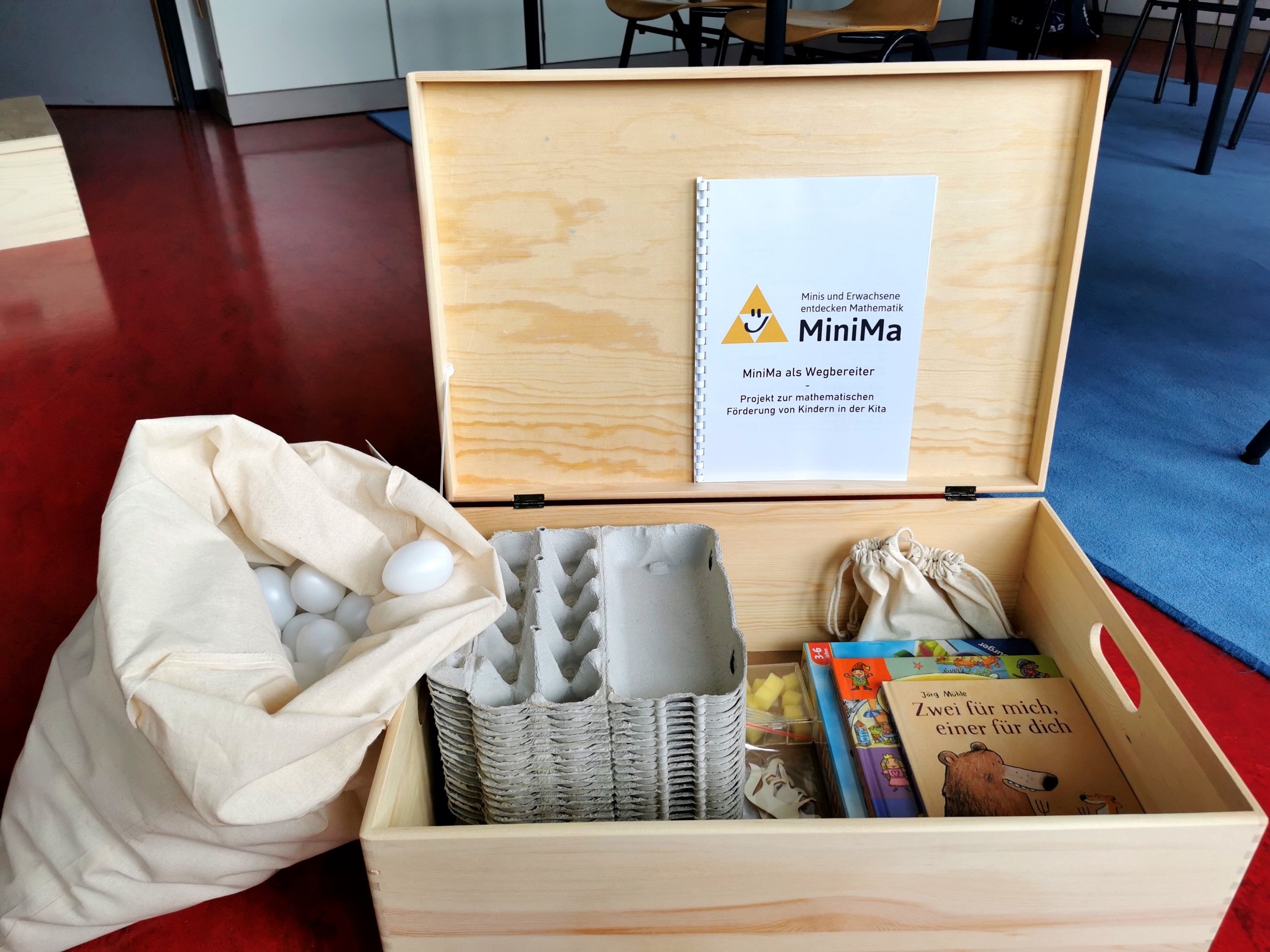 Eine helle Holzkiste mit Materialien zur Mathematikförderung wie Eierschachteln, daneben ein weißer Sack gefüllt mit Plastikeiern