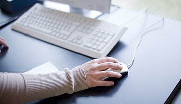 Ausschnitt eines Arms, dessen Hand auf einer Computermaus aufliegt. Davor ist eine Computertastatur der Bibliothek zu sehen. Die Bibliothek befindet sich im Gebäude 2 der PH Karlsruhe.