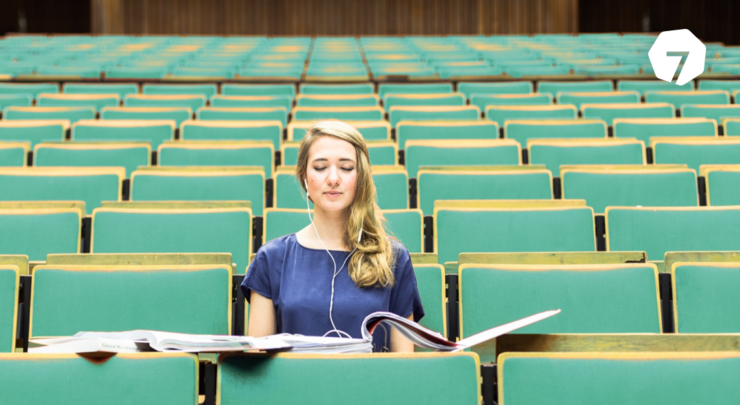 Eine Studentin sitzt alleine mit geschlossenen Augen in einem großen Hörsaal. Sie hat die Augen geschlossen und Kopfhörer auf. Vor ihr liegen aufgeschlagene Ordner.