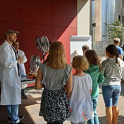 Ein Student im weißen Kittel erklärt Kindern (sie sind von hinten zu sehen) im Freien ein natruwissenschaftliches Experiment - im Hintergrund ist u.a. ein Venntilator zu sehen - im Rahmen des Ferienprogramms RELAX