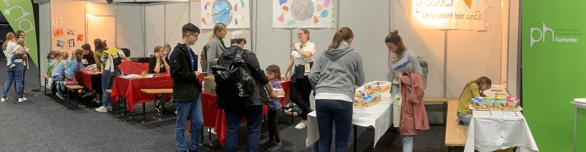 Reihe der Ausstellertische der PHKA bei den Science DAys für Kinder mit roten Tischdecken. Flankiert wird es von aufstelern mit dem PHKA Logo weiß auf Grün. Um die Tische herum sitzen und stehen KInder und Studierende.