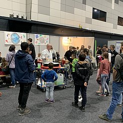 Blick in die Veranstaltungshalle des Europaparks auf den Stand der PHKA, im Hintergrund Tische und Plakate, im vorfeld führt ein Mann Experimente vor und es scharen sich Kinder und Erwachsene um ihn.