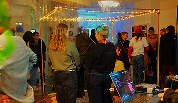 Situation auf der Kunstausstellung mit ein paar Personen, die eine Installation bettrachten, und weiteren im Hintergrund.