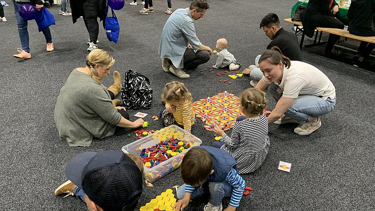 PHKA auf den Science Days für Kinder 2023 6 Kinder, Eltern und PHKA Studierende kauern, knien und sitzen auf dem Boden und legen mit bunten geometrischen Formen aus Holz Bandornamente und Parkettmuster