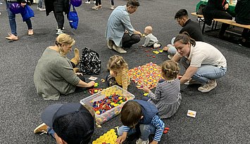 Kinder, Eltern und PHKA Studierende kauern, knien und sitzen auf dem Boden und legen mit bunten geometrischen Formen aus Holz Bandornamente und Parkettmuster
