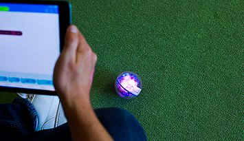 Eine jugendliche Person von hinten, die einTablet in den Händen hält, mit dem sie das auf dem grünen Boden zu sehende Sphero steuert.