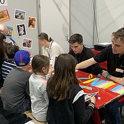 Bei den Science DAys für Kinder erklärt ein Mathematikstudent Kindern, wie aus verschlungenen Papierstreifen Bilderrahmen erstellt werden können. Die Materialien sind auf dem Tisch zu sehen.
