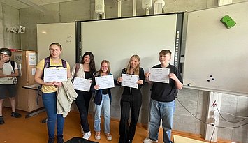Fünf jugendliche Schüler:innen zeigen stolz ihre durch einen Hackathon des Projekts EntreCo Think erworbenen Zertifikate vor einem Whiteboard.