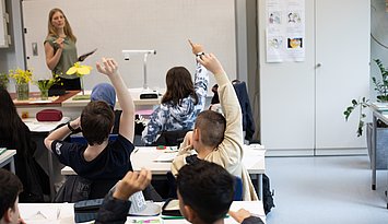 Eine Lehrerin steht vor einer Schulklasse. Dei Schülerinnen und Schüler melden sich teilweise und sind von hinten zu sehen.