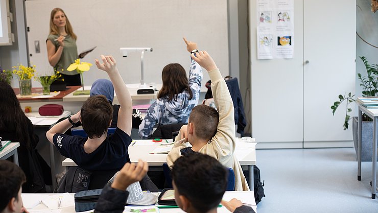 Unterrichtsszene Eine Lehrerin steht vor einer Schulklasse. Dei Schülerinnen und Schüler melden sich teilweise und sind von hinten zu sehen.