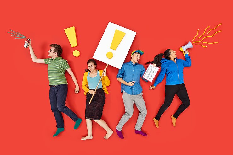 4 Personen auf rotem Hintergrun, die mit einem Megafon, Plakaten mit Ausrufezeichen und einer Spraydose Aufmerksamkeit erzeugen