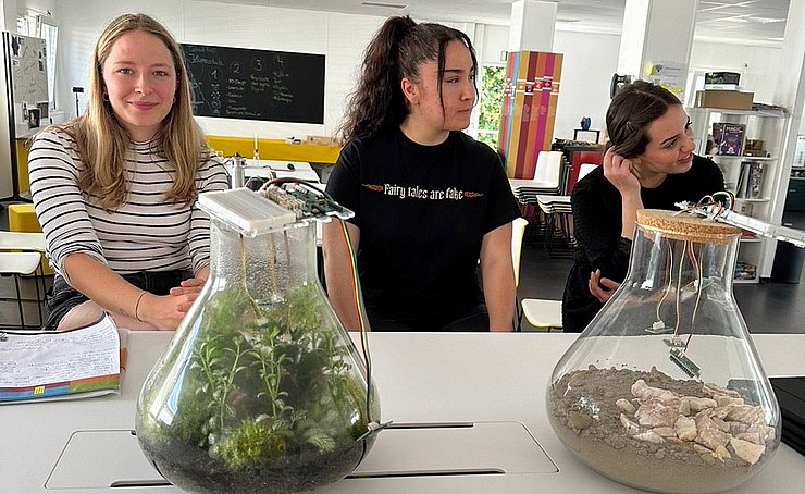 Vorbereitung Kiwanis Sommerschule 2023_2 Drei Studentinnen sitzen an einem Tisch in einem Labor. Vor Ihnen zwei Glasgefäße mit Erde und Pflanzen darin. Das Thema ist Nachhaltigkeit.