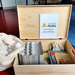 Eine helle Holzkiste mit Materialien zur Mathematikförderung wie Eierschachteln, daneben ein weißer Sack gefüllt mit Plastikeiern