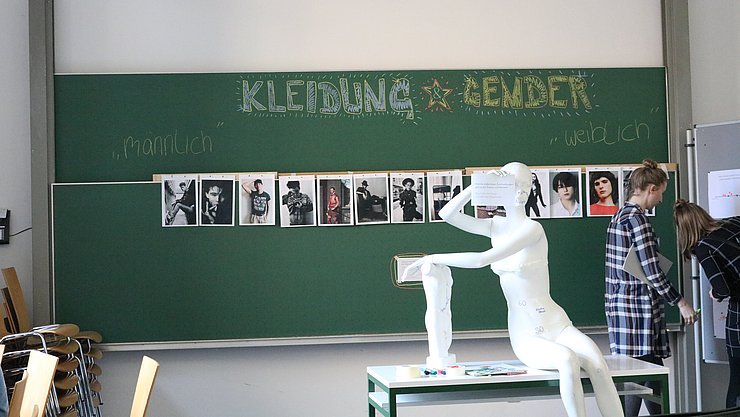 Bild der Station der Soziologie mit weißer Puppe zum mit Nachrichten Bekleben und Tafel mit Bildern zu Kleidung genderkritisch betrachtet