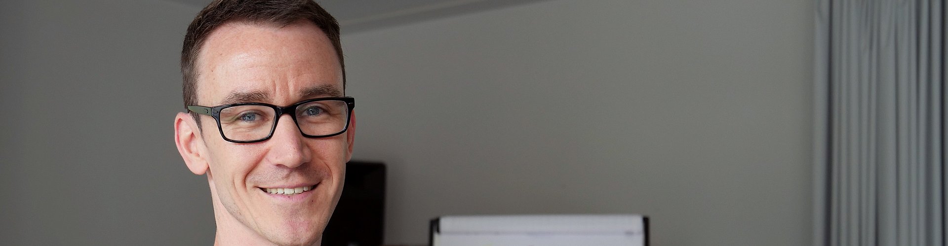 Eine Person steht im Vordergrund und blickt in Kamera. Im Hintergrund arbeiten zwei Personen an einer Mindmap.