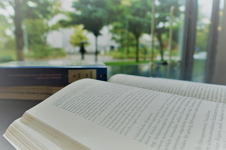 Das Foto zeigt eine Nahaufnahme von einem Schreibtisch der Hochschulbibliothek mit Blick ins Grüne. Auf dem Bild sieht man ein aufgeschlagenes Buch, im Hintergrund stapeln sich weitere Bücher.