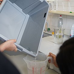 Zu sehen ist das übergelaufene Wasser in einem Behälter, welcher nun in einen Messbecher umgefüllt wird.