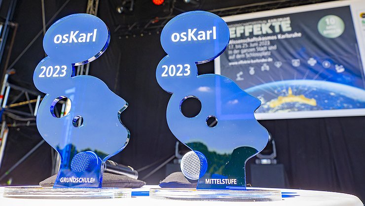 osKarl-Verleihung EFFEKTE 2023 Jürgen Rösner Zwei osKarl Pokale in Blauauf der Festivalbohne, im Hintergrund eine Leinwand mit dem EFFEKTE-Plakat