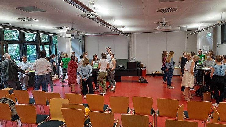 In einem Hörsaal der PHKA mit rotem Boden stehen verteilt an Stehtischen sich unterhaltende Personen.