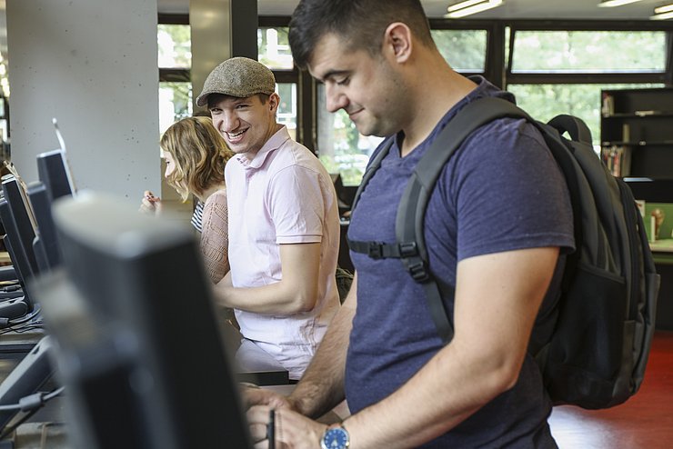 Ein Student, von der Seite zu sehen, sucht ein Buch an einem Computer mithilfe eines für die Medienrecherche ausgewiesenen Programm. Neben ihm steht ein weiterer lächelnder Student. Sie befinden sich in der Bibliothek im Gebäude 2 der PH Karlsruhe.