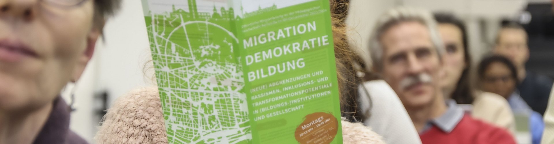 Das Publikum der Ringvorlesung Migration-Demokratie-Bildung: Eine Zuschauerin hält die Broschüre der Ringvorlesung in den Händen.