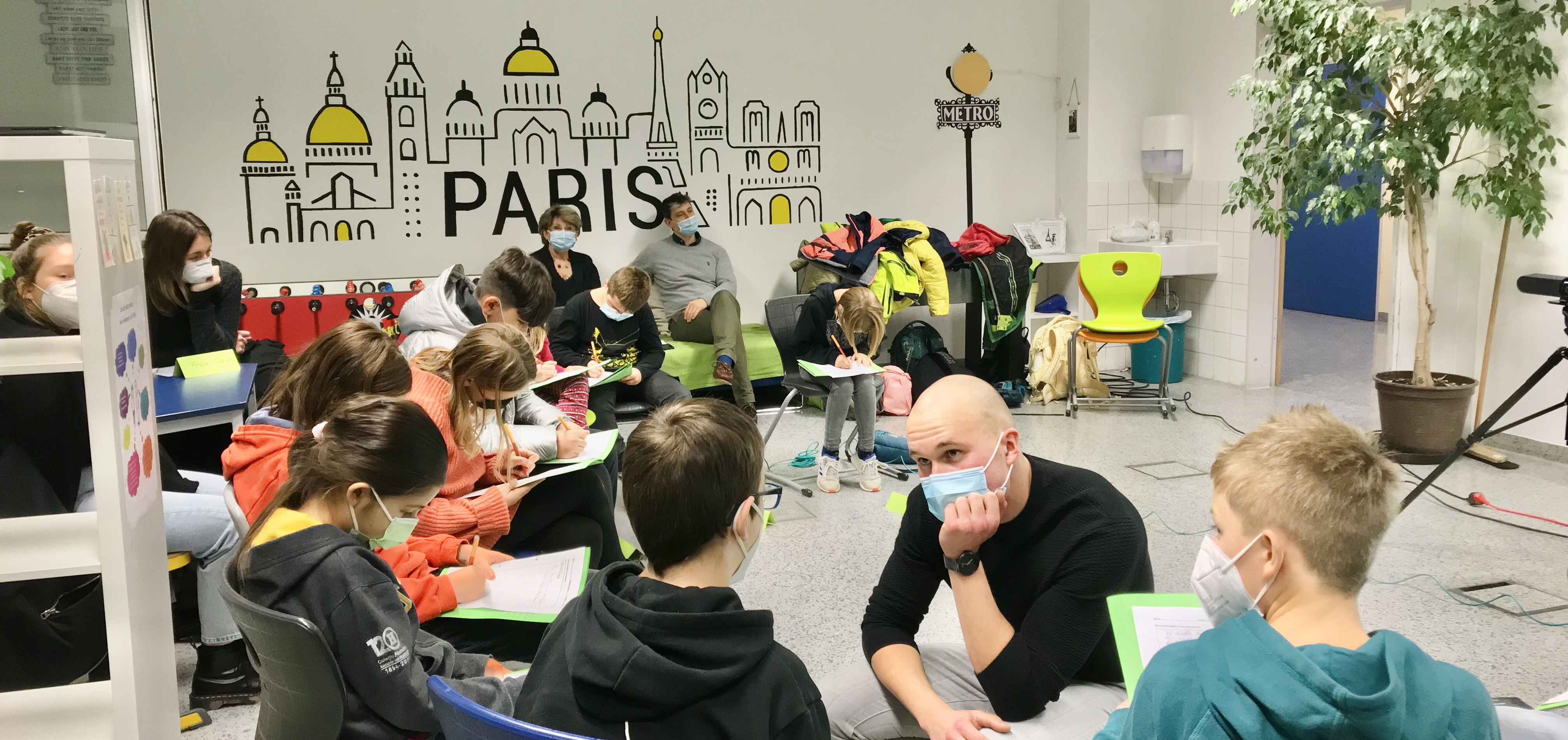 In einem hellen Gruam der Brettener Gesamtschule sitzen Kinder im Kreis und diskutieren mit einem PH Studenten über Philosophie. auf der wand im Hintetrgrund ist schematisch ein Häuserzug mit dem Schriftzug "Paris" zu sehen.
